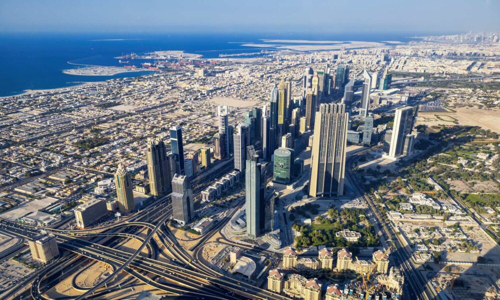 Aerial view of Dubai's Financial District skyline with modern skyscrapers, highways, and coastal development extending to the Persian Gulf.
