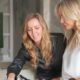 Two blonde women reviewing design materials at Blush International studio with green lamp and white flowers in background.