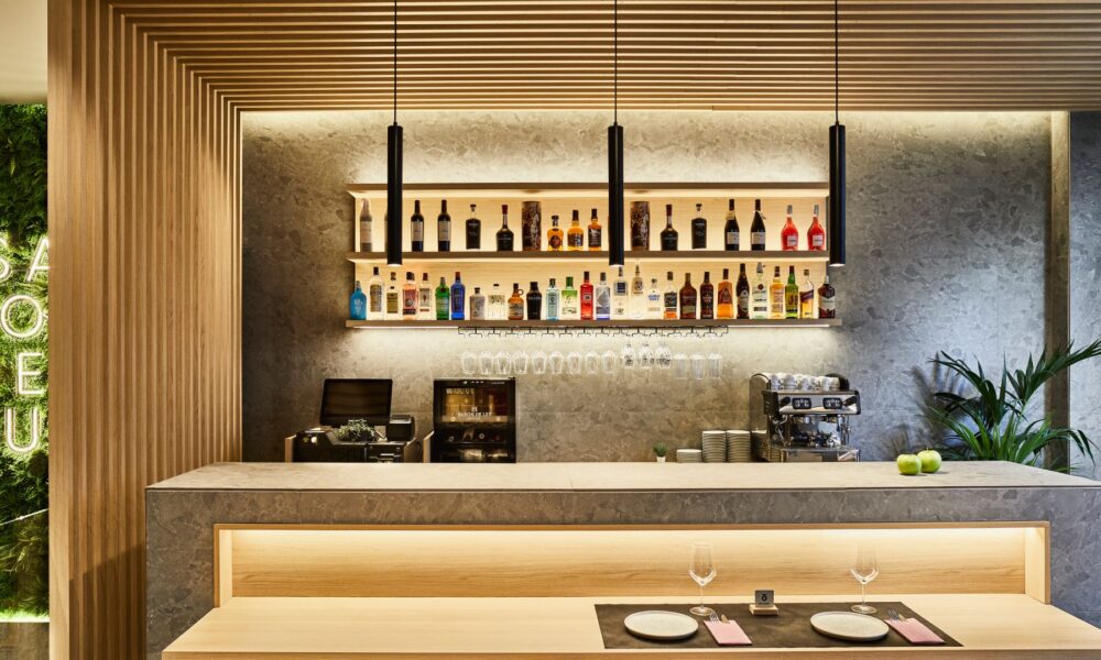 Modern restaurant bar featuring large-format ceramic slabs, wood slat ceiling, illuminated shelving with bottles, and marble-effect surfaces.