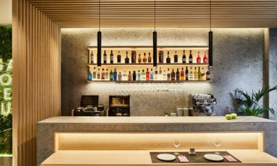 Modern restaurant bar featuring large-format ceramic slabs, wood slat ceiling, illuminated shelving with bottles, and marble-effect surfaces.