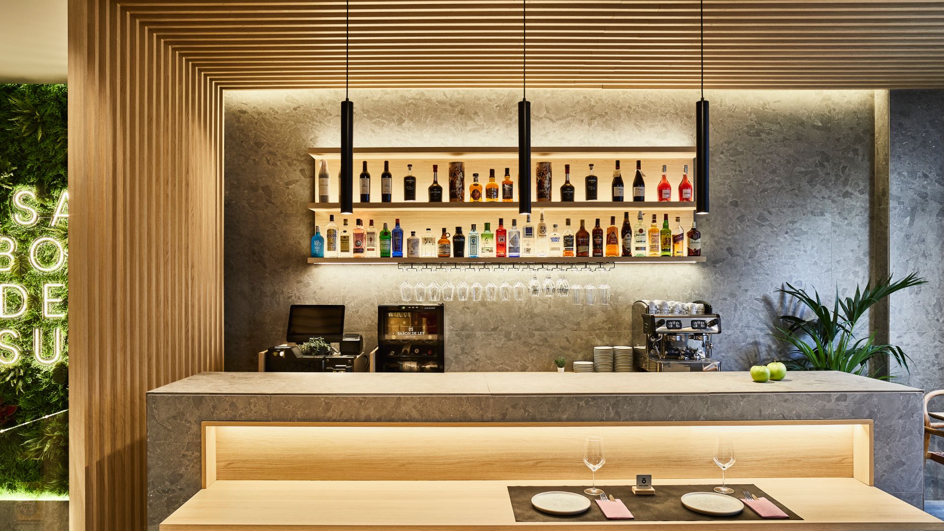 Modern restaurant bar featuring large-format ceramic slabs, wood slat ceiling, illuminated shelving with bottles, and marble-effect surfaces.