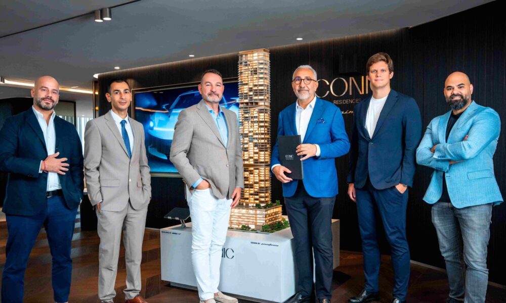 Six business executives in suits standing around illuminated ICONIC Residences tower model in modern showroom with blue lighting and branded wall display.