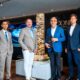Six business executives in suits standing around illuminated ICONIC Residences tower model in modern showroom with blue lighting and branded wall display.