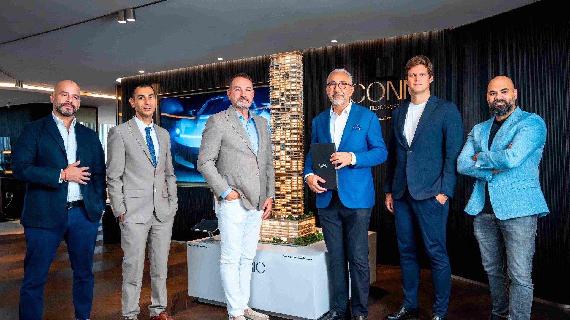 Six business executives in suits standing around illuminated ICONIC Residences tower model in modern showroom with blue lighting and branded wall display.