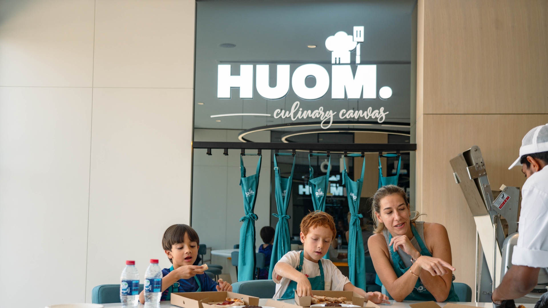 Mother and two children cooking together at HUOM Culinary Canvas cooking studio with chef instructor in modern kitchen setting.