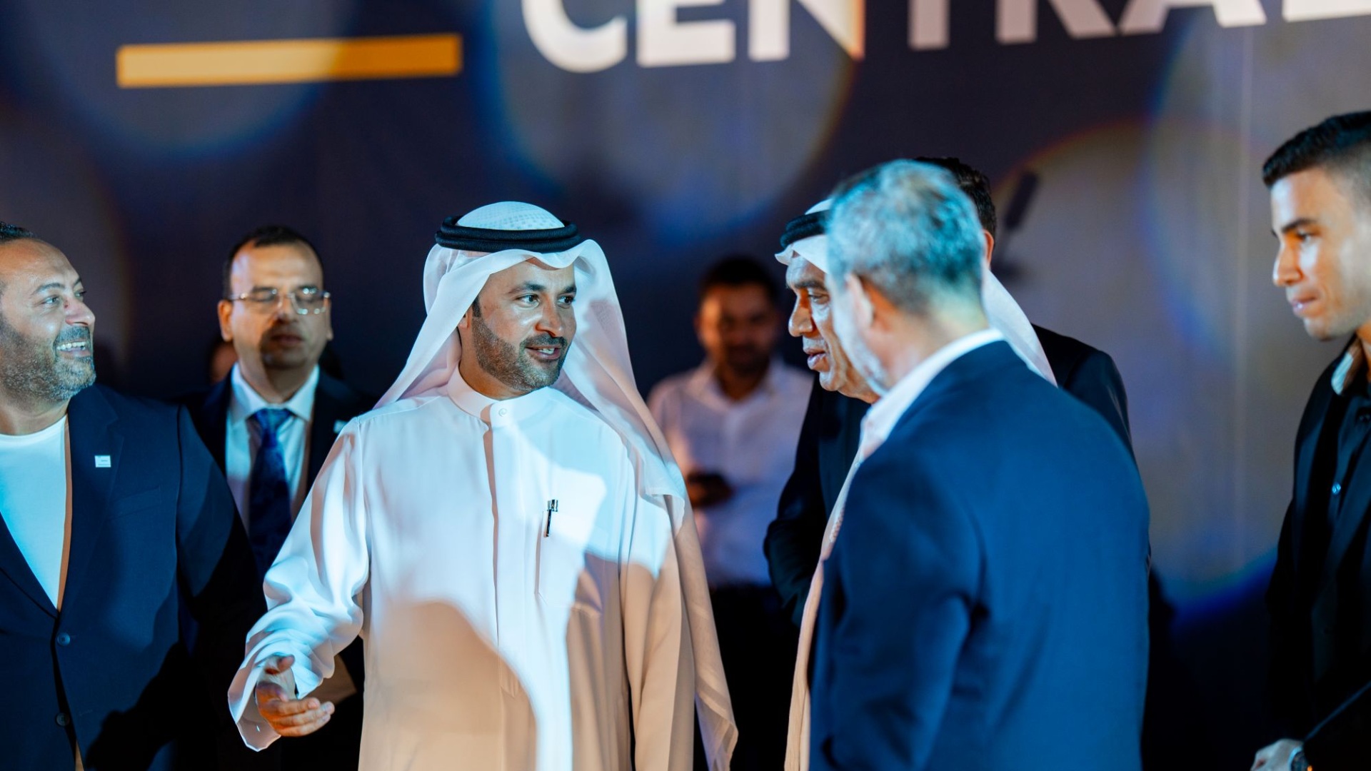 Group of business leaders in formal attire conversing at an event, with one man in traditional Emirati dress at the center