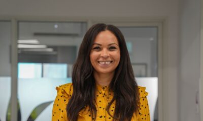 Professional woman in yellow polka dot blouse smiling in modern office setting with contemporary kitchen design in background.