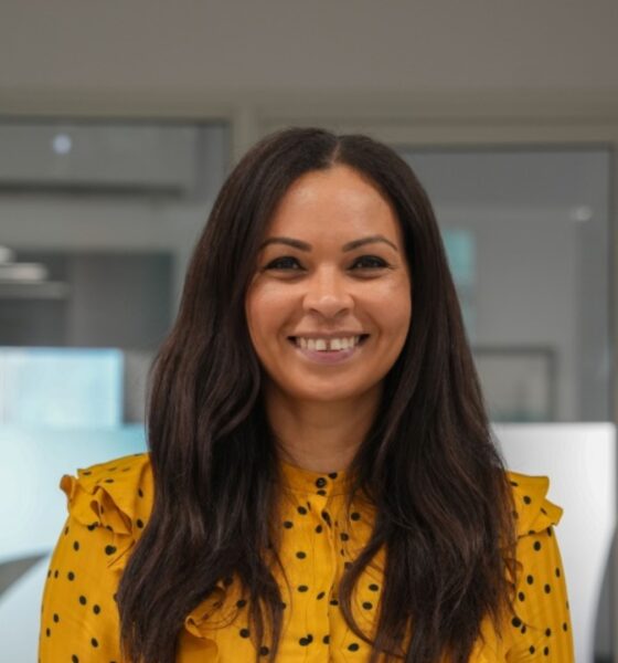 Professional woman in yellow polka dot blouse smiling in modern office setting with contemporary kitchen design in background.