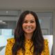Professional woman in yellow polka dot blouse smiling in modern office setting with contemporary kitchen design in background.
