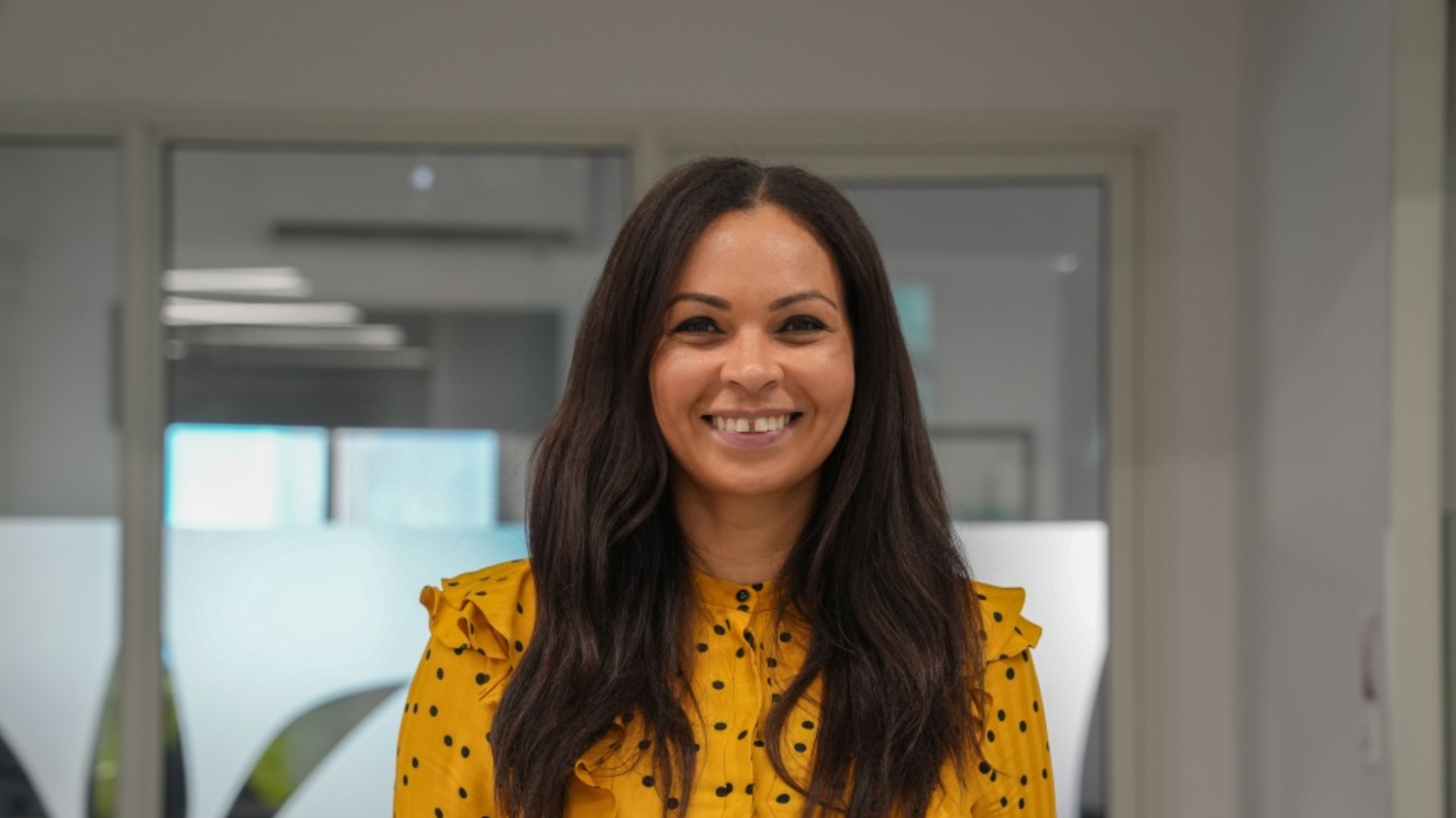 Professional woman in yellow polka dot blouse smiling in modern office setting with contemporary kitchen design in background.