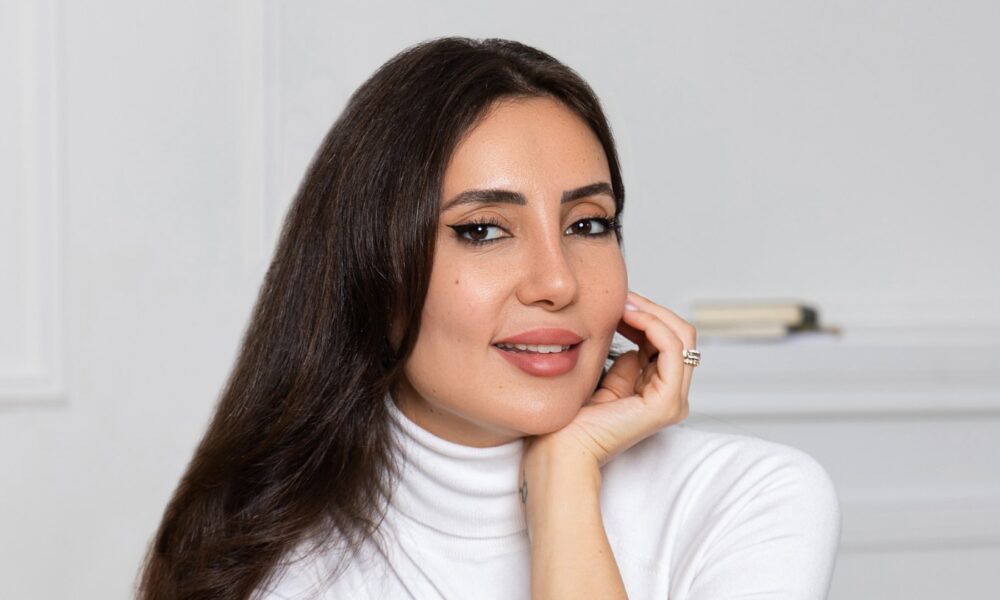 Professional woman in white turtleneck with long dark hair smiling at camera in bright minimalist office setting.