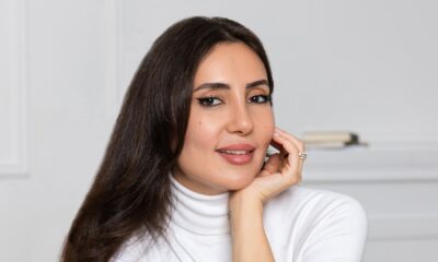 Professional woman in white turtleneck with long dark hair smiling at camera in bright minimalist office setting.
