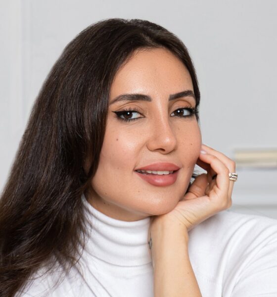 Professional woman in white turtleneck with long dark hair smiling at camera in bright minimalist office setting.