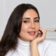 Professional woman in white turtleneck with long dark hair smiling at camera in bright minimalist office setting.