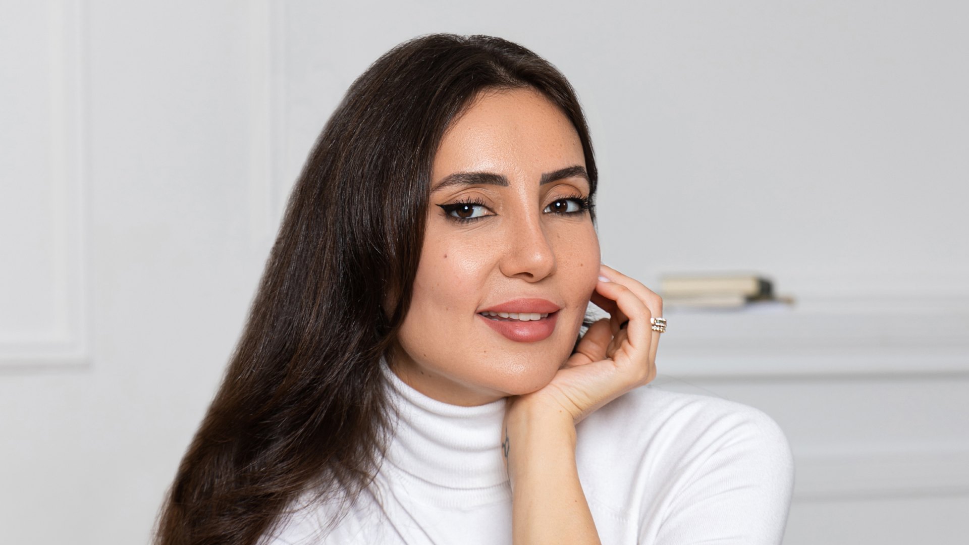 Professional woman in white turtleneck with long dark hair smiling at camera in bright minimalist office setting.
