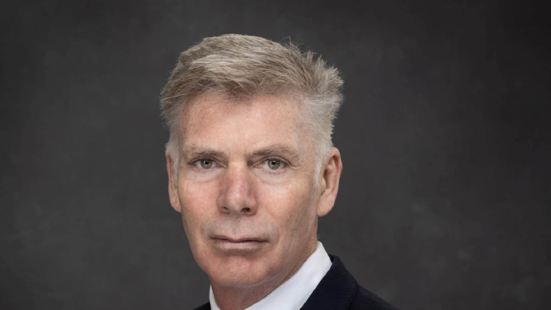 Professional headshot of a businessman in dark suit and white shirt against gray background
