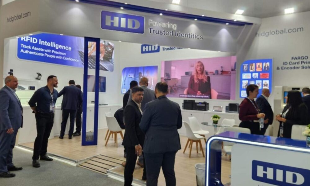 HID Global exhibition booth at trade show featuring RFID Intelligence and ID card printing solutions, with attendees in business attire viewing displays and a video presentation