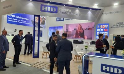 HID Global exhibition booth at trade show featuring RFID Intelligence and ID card printing solutions, with attendees in business attire viewing displays and a video presentation