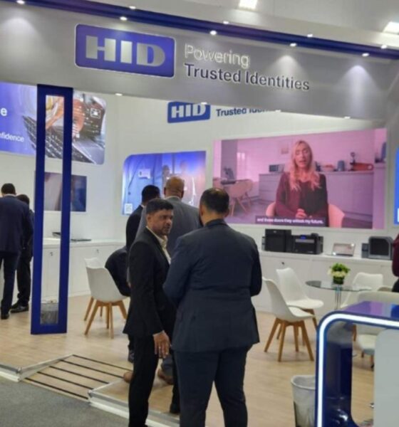 HID Global exhibition booth at trade show featuring RFID Intelligence and ID card printing solutions, with attendees in business attire viewing displays and a video presentation
