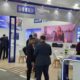 HID Global exhibition booth at trade show featuring RFID Intelligence and ID card printing solutions, with attendees in business attire viewing displays and a video presentation