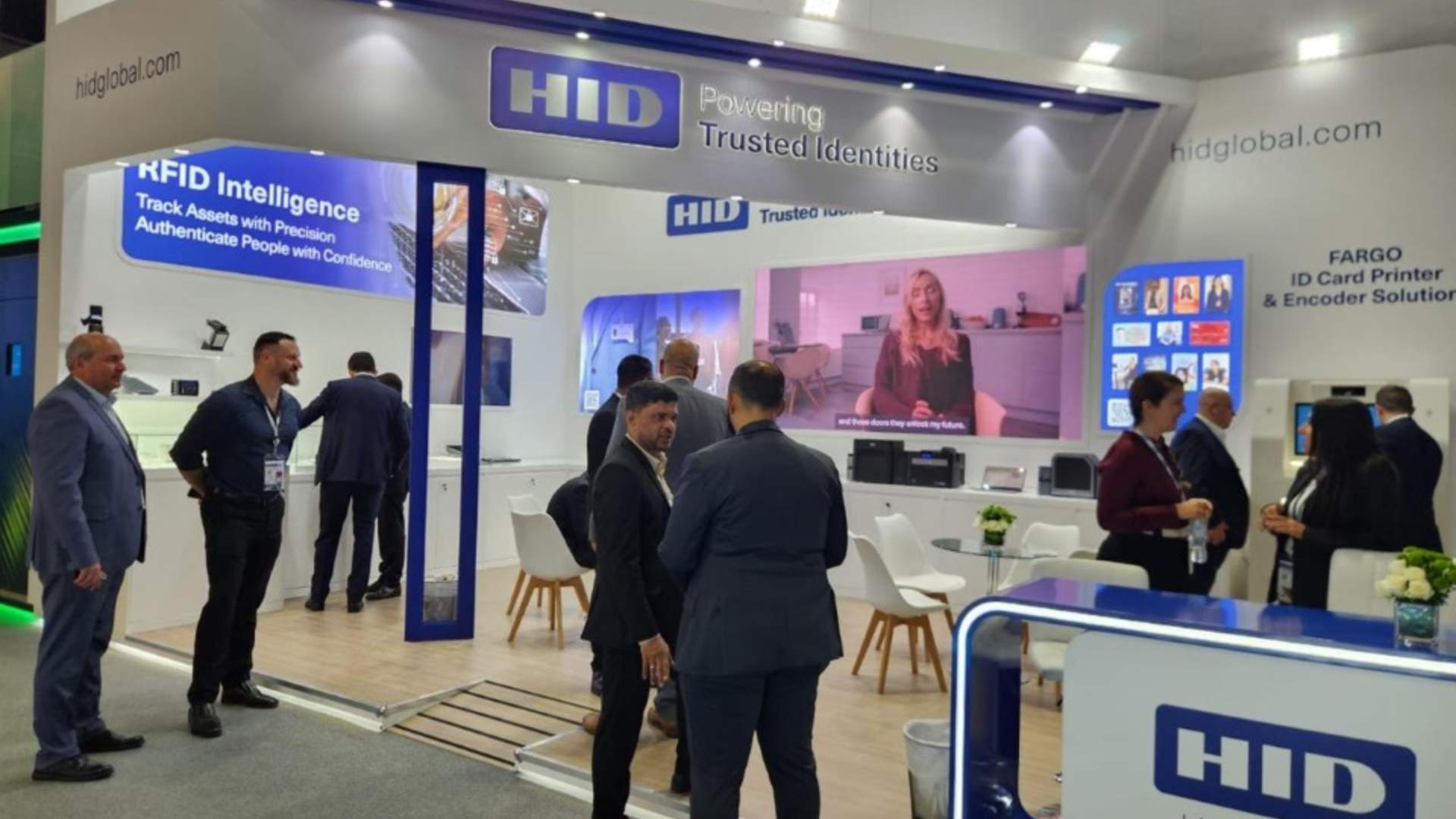HID Global exhibition booth at trade show featuring RFID Intelligence and ID card printing solutions, with attendees in business attire viewing displays and a video presentation