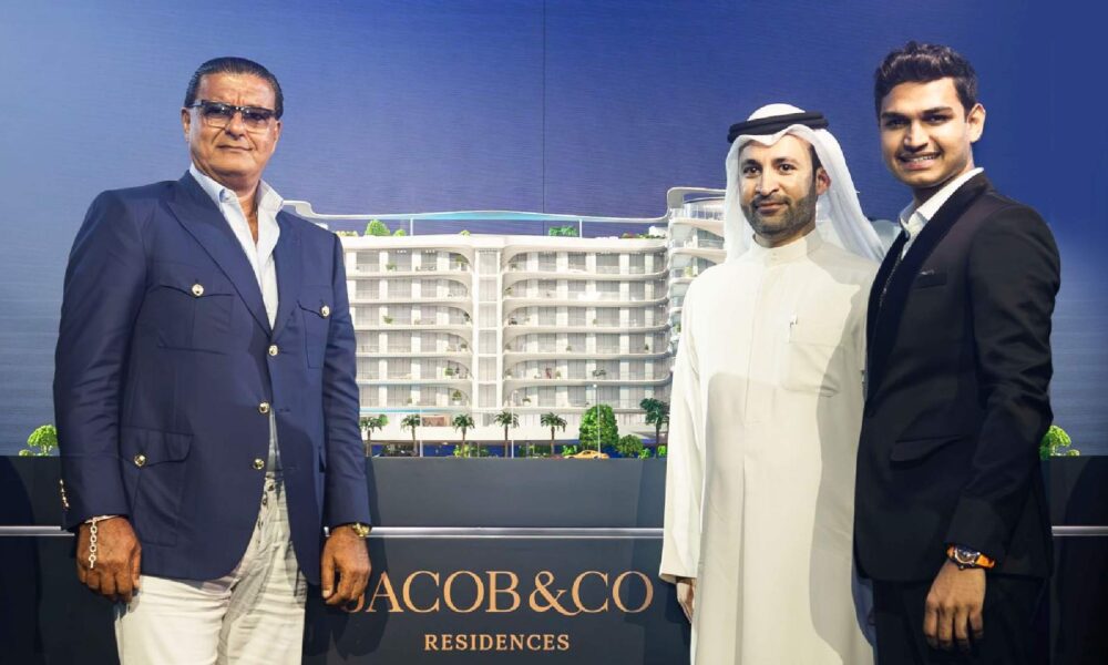 Three business professionals posing with an architectural model of Jacob & Co Residences, a modern multi-story residential building with palm trees and rooftop greenery