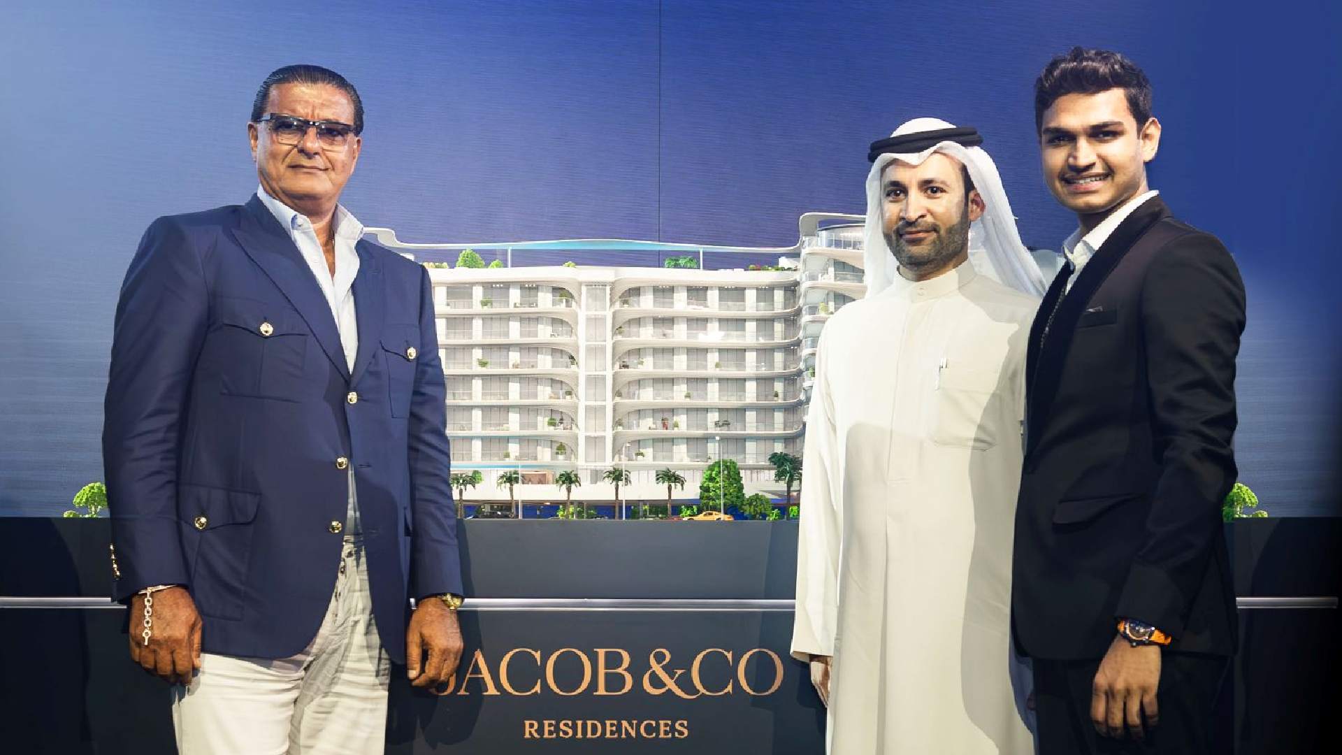 Three business professionals posing with an architectural model of Jacob & Co Residences, a modern multi-story residential building with palm trees and rooftop greenery