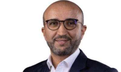 Professional headshot of smiling businessman wearing round glasses and dark suit against white background