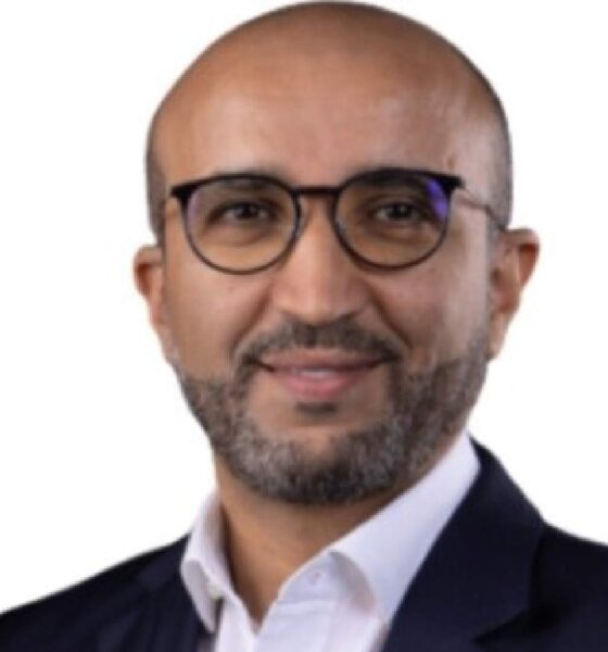 Professional headshot of smiling businessman wearing round glasses and dark suit against white background