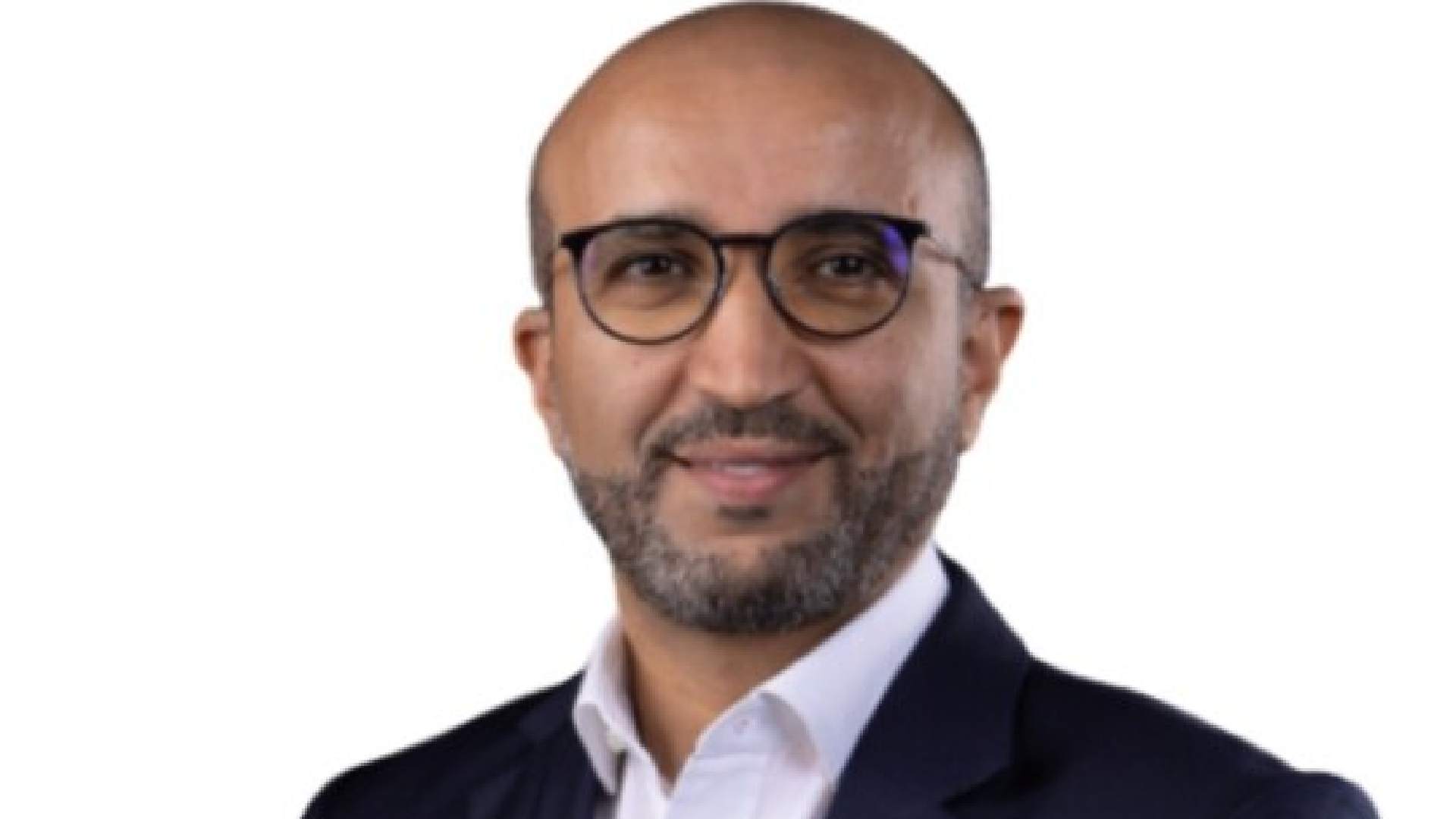 Professional headshot of smiling businessman wearing round glasses and dark suit against white background
