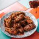Hand holding glazed chicken wing dripping with sauce over plate of barbecue wings on geometric patterned plate