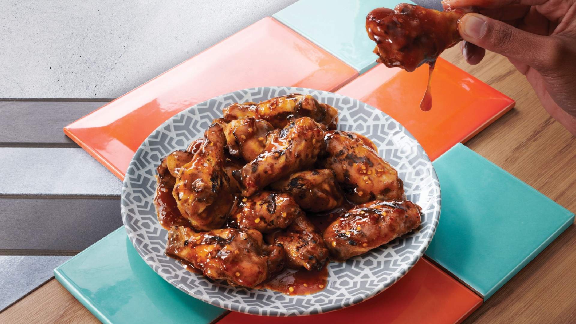 Hand holding glazed chicken wing dripping with sauce over plate of barbecue wings on geometric patterned plate