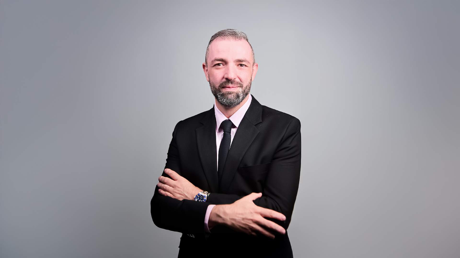 Professional businessman in black suit with arms crossed standing against gray background
