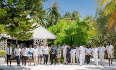 Diverse group of resort staff in white and casual attire standing together at tropical beachfront property with palm trees and thatched roof villas