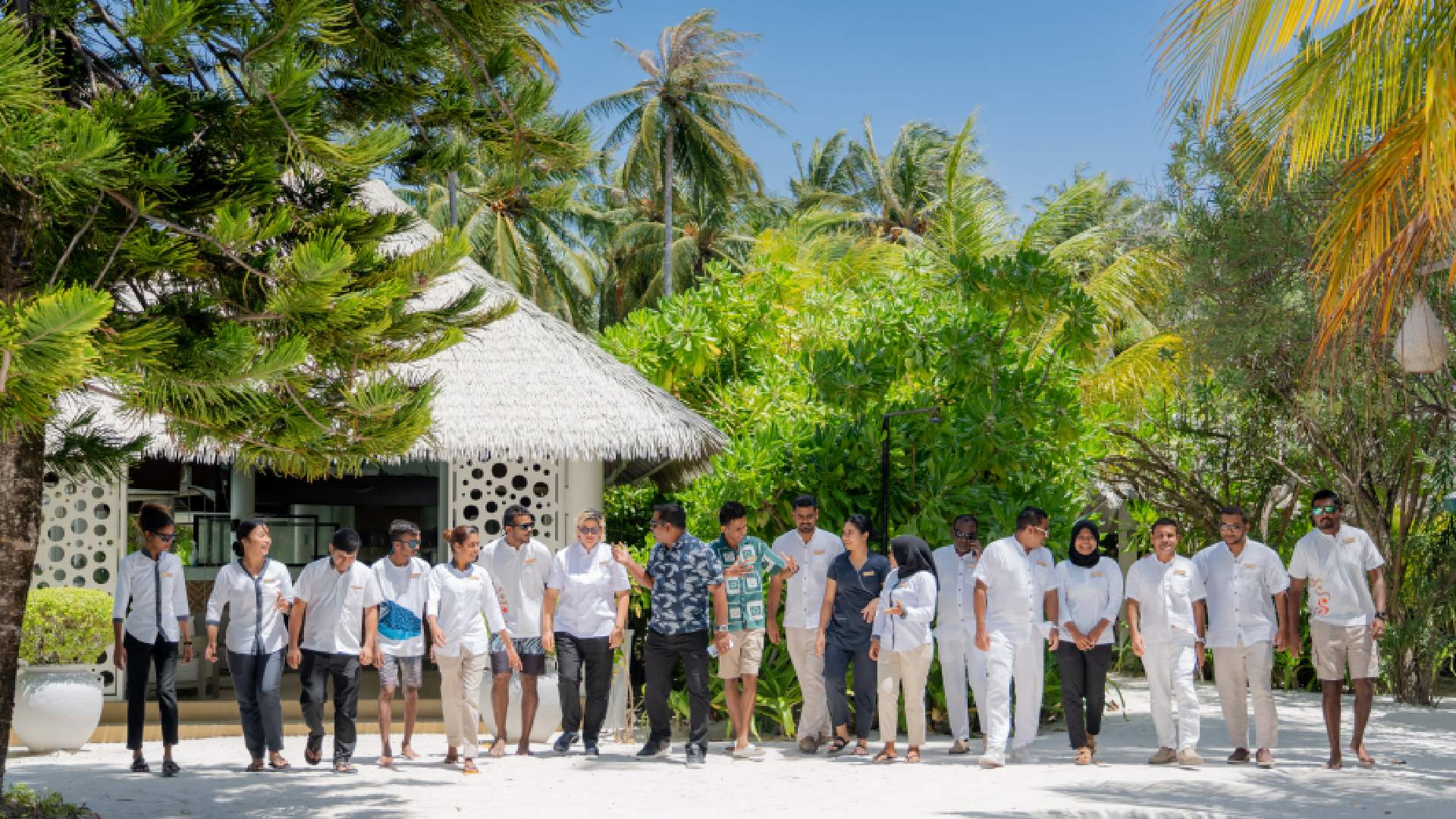 Diverse group of resort staff in white and casual attire standing together at tropical beachfront property with palm trees and thatched roof villas