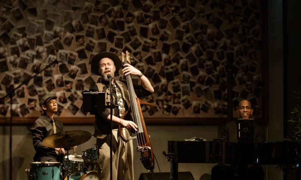 Three-piece jazz trio performing live at Cucina The Palm during Jazz and Juice Nights, featuring upright bass, piano, and drums against decorative wall backdrop.