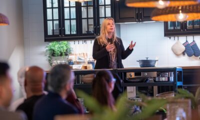 A blonde woman in a black jacket is speaking to a seated audience in what appears to be an IKEA kitchen showroom, with dark cabinets, a white subway tile backsplash, and hanging wicker-style lamps above.