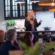 A blonde woman in a black jacket is speaking to a seated audience in what appears to be an IKEA kitchen showroom, with dark cabinets, a white subway tile backsplash, and hanging wicker-style lamps above.