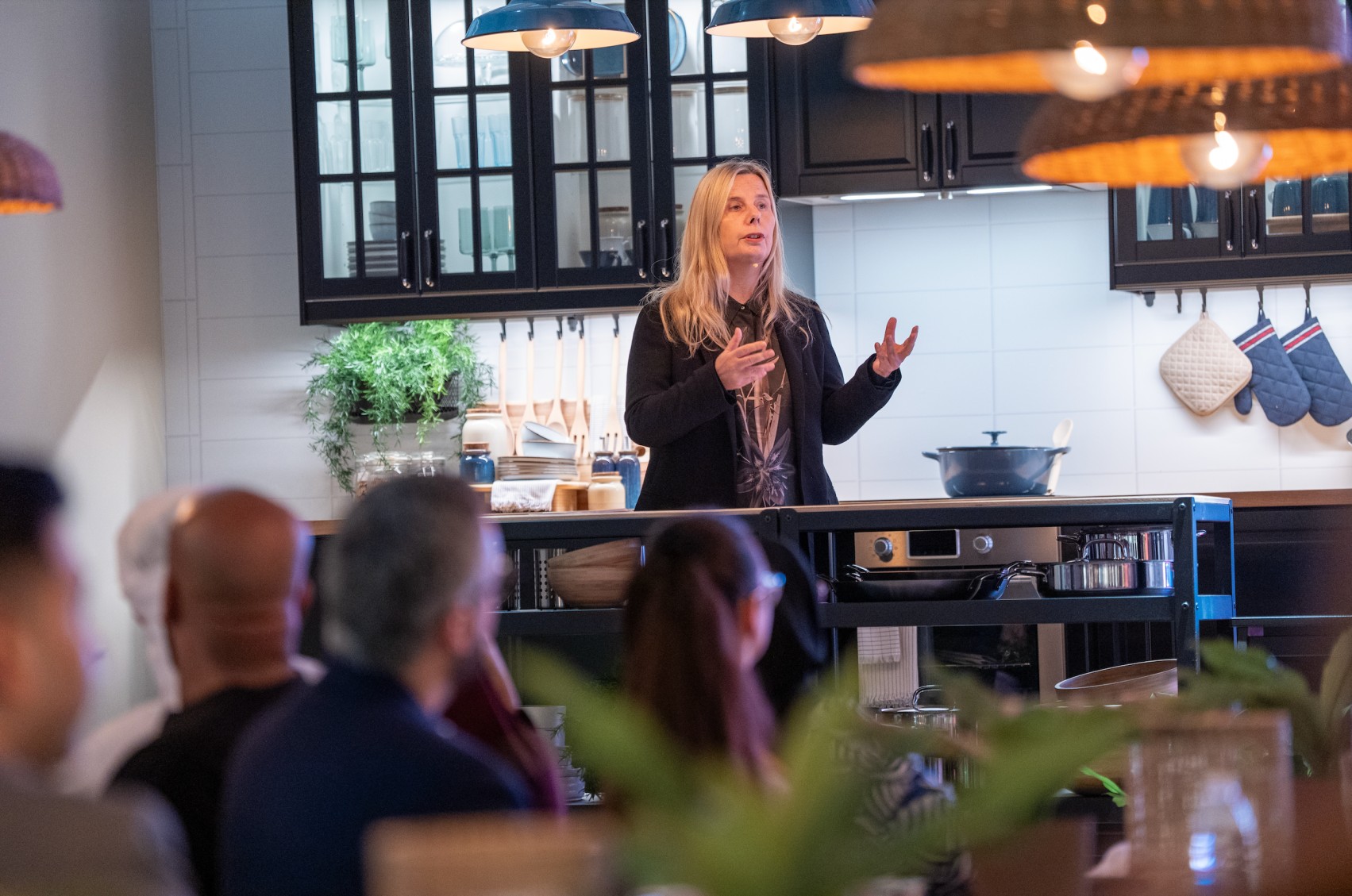A blonde woman in a black jacket is speaking to a seated audience in what appears to be an IKEA kitchen showroom, with dark cabinets, a white subway tile backsplash, and hanging wicker-style lamps above.