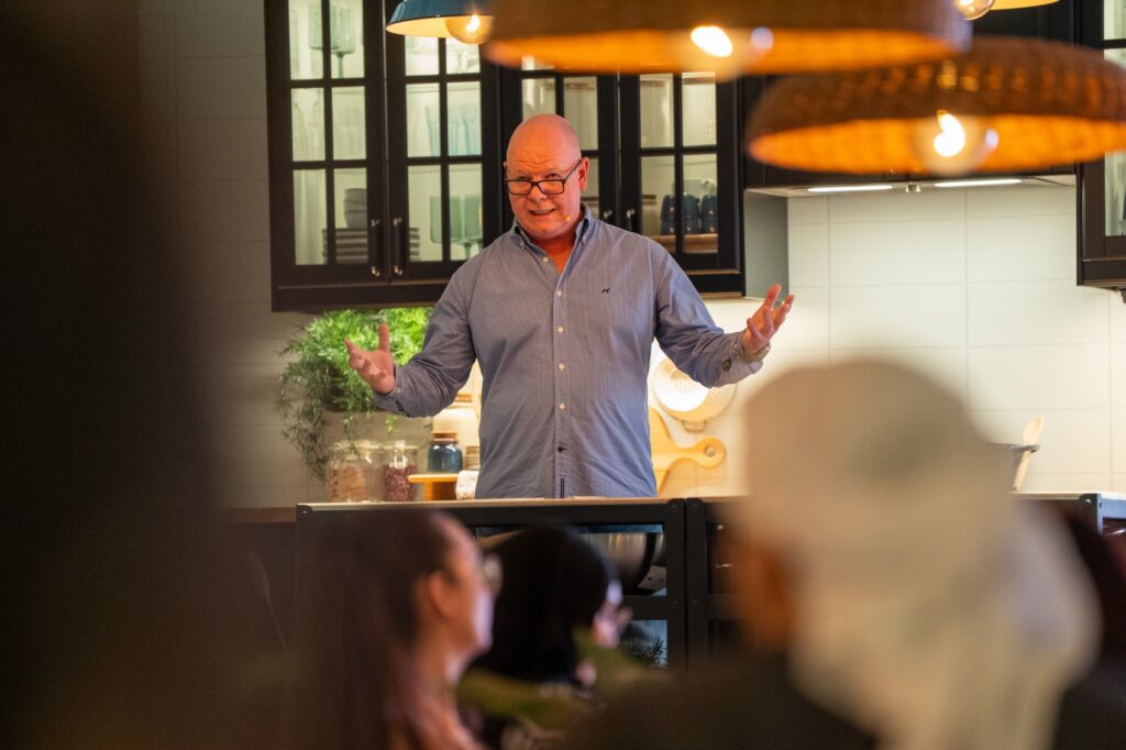 A bald man wearing glasses and a light blue checkered shirt is speaking enthusiastically with his hands outstretched to an audience, positioned in an IKEA kitchen showroom with dark cabinets and wicker lamps.