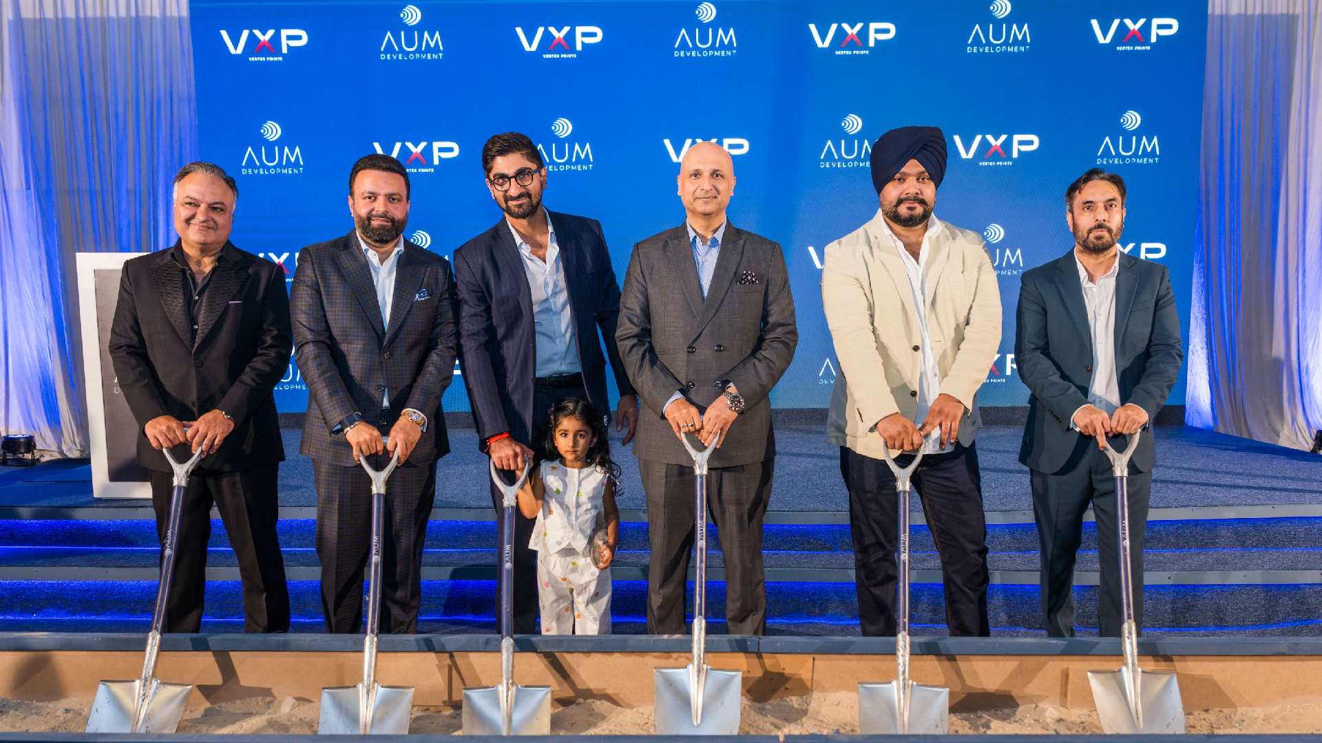 Business executives at groundbreaking ceremony holding silver shovels in front of VXP and AUM Development branded backdrop