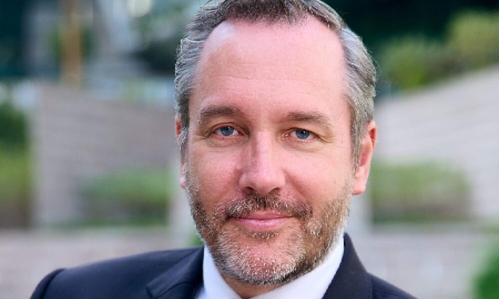 Professional headshot of a businessman with blue eyes and grey-brown beard wearing a dark suit against a blurred outdoor background