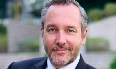 Professional headshot of a businessman with blue eyes and grey-brown beard wearing a dark suit against a blurred outdoor background