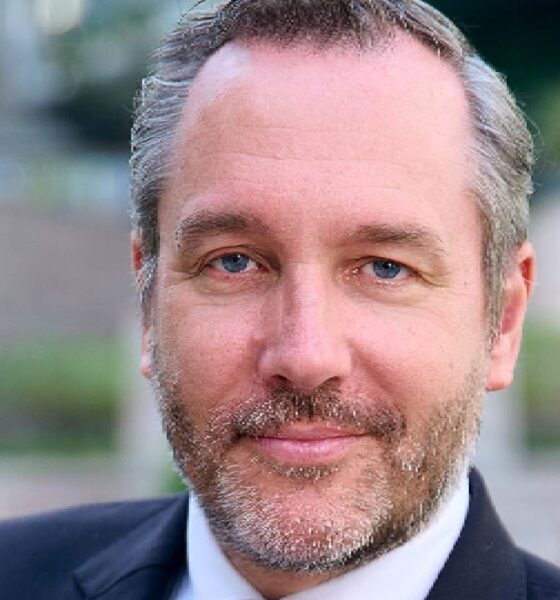 Professional headshot of a businessman with blue eyes and grey-brown beard wearing a dark suit against a blurred outdoor background
