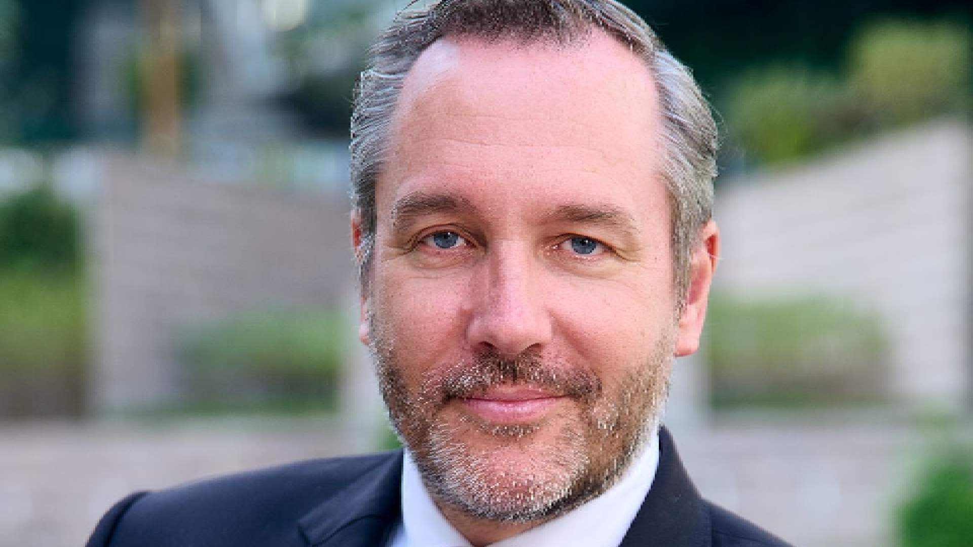 Professional headshot of a businessman with blue eyes and grey-brown beard wearing a dark suit against a blurred outdoor background