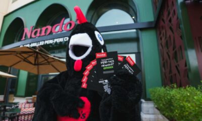 Barci, Nando's black and red rooster mascot, holding scratch-and-win cards outside Nando's JBR restaurant in Dubai