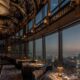 Luxury restaurant interior with ornate decorative ceiling, floor-to-ceiling windows, and panoramic Dubai cityscape views at twilight