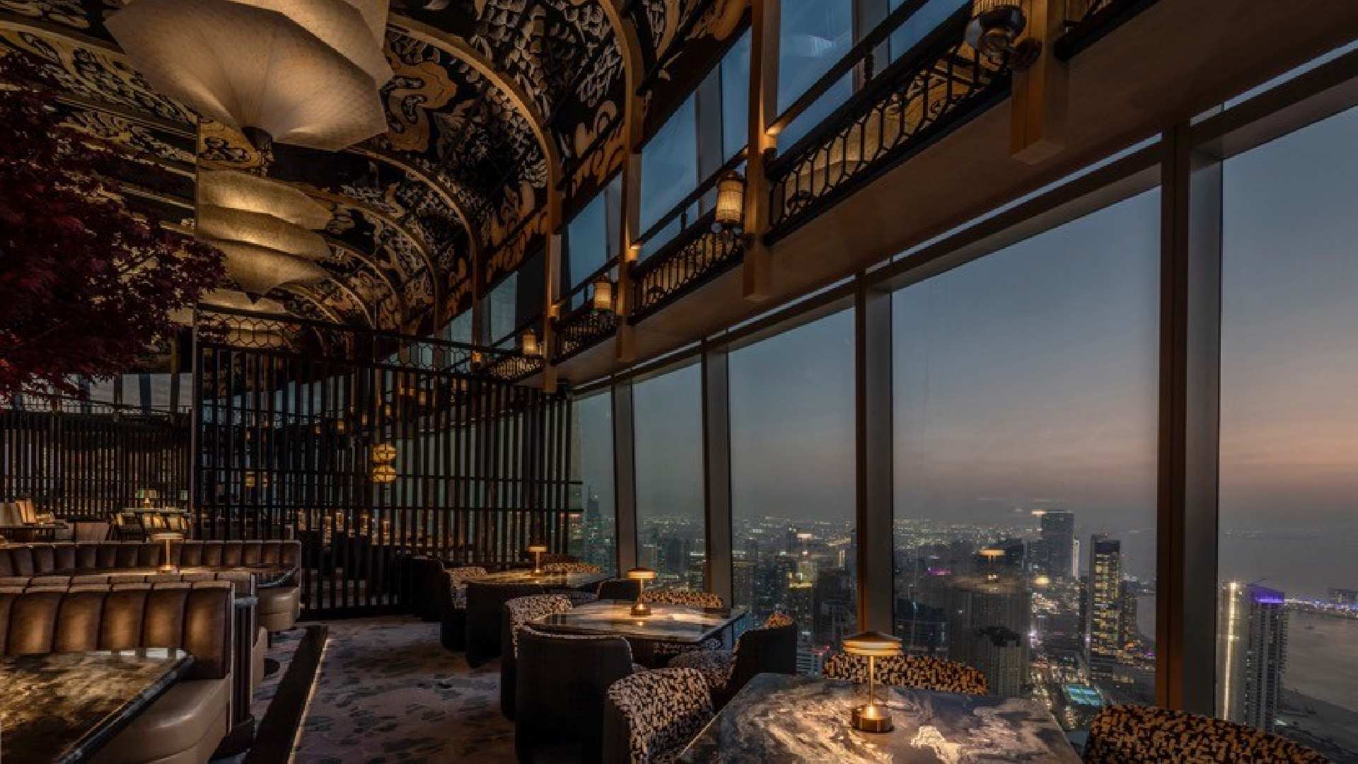 Luxury restaurant interior with ornate decorative ceiling, floor-to-ceiling windows, and panoramic Dubai cityscape views at twilight