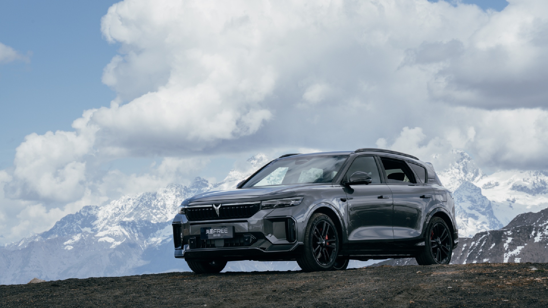 Dark gray SUV with black wheels positioned on mountain terrain with snow-capped peaks and cloudy sky in background