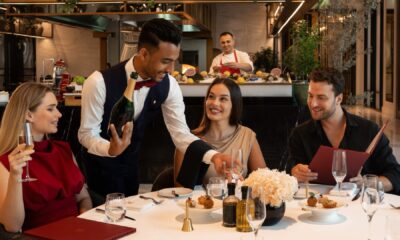 A waiter in a formal vest is presenting a bottle of champagne to a smiling woman seated at a white-clothed dinner table with two other diners in an upscale restaurant.